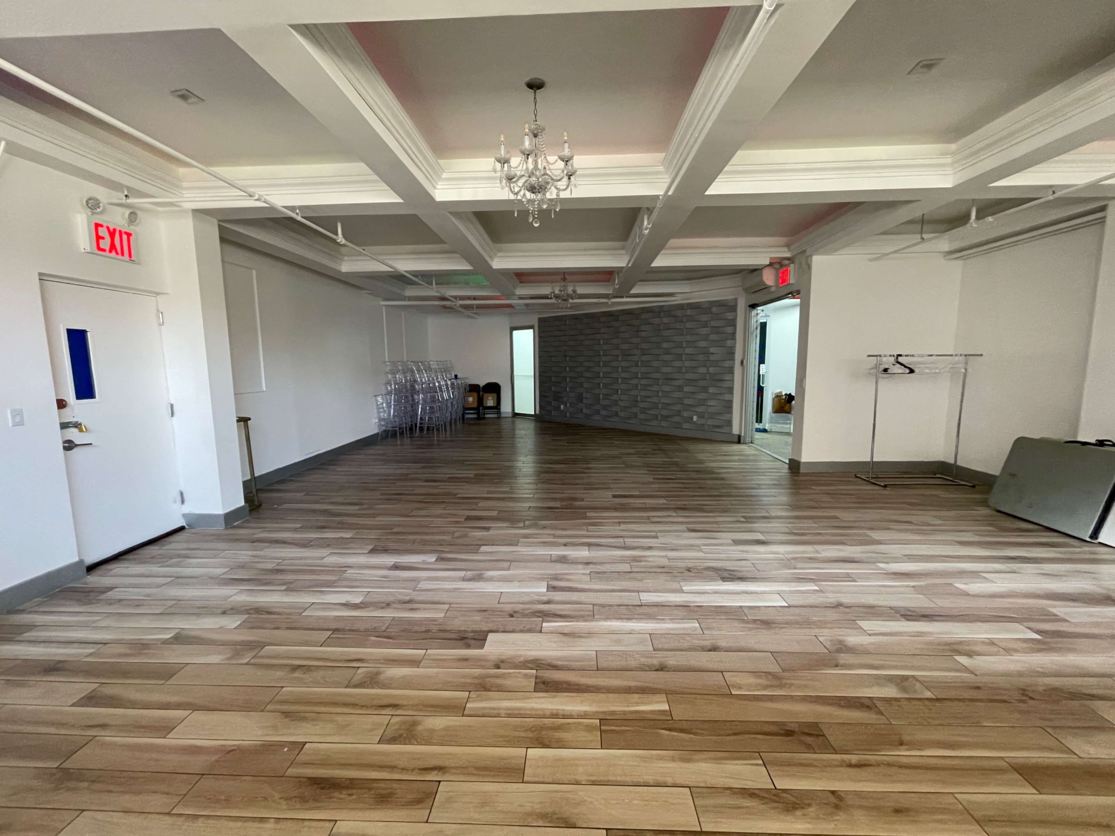 A spacious, empty room with wooden flooring and decorative ceiling beams, featuring a chandelier and exit signs.