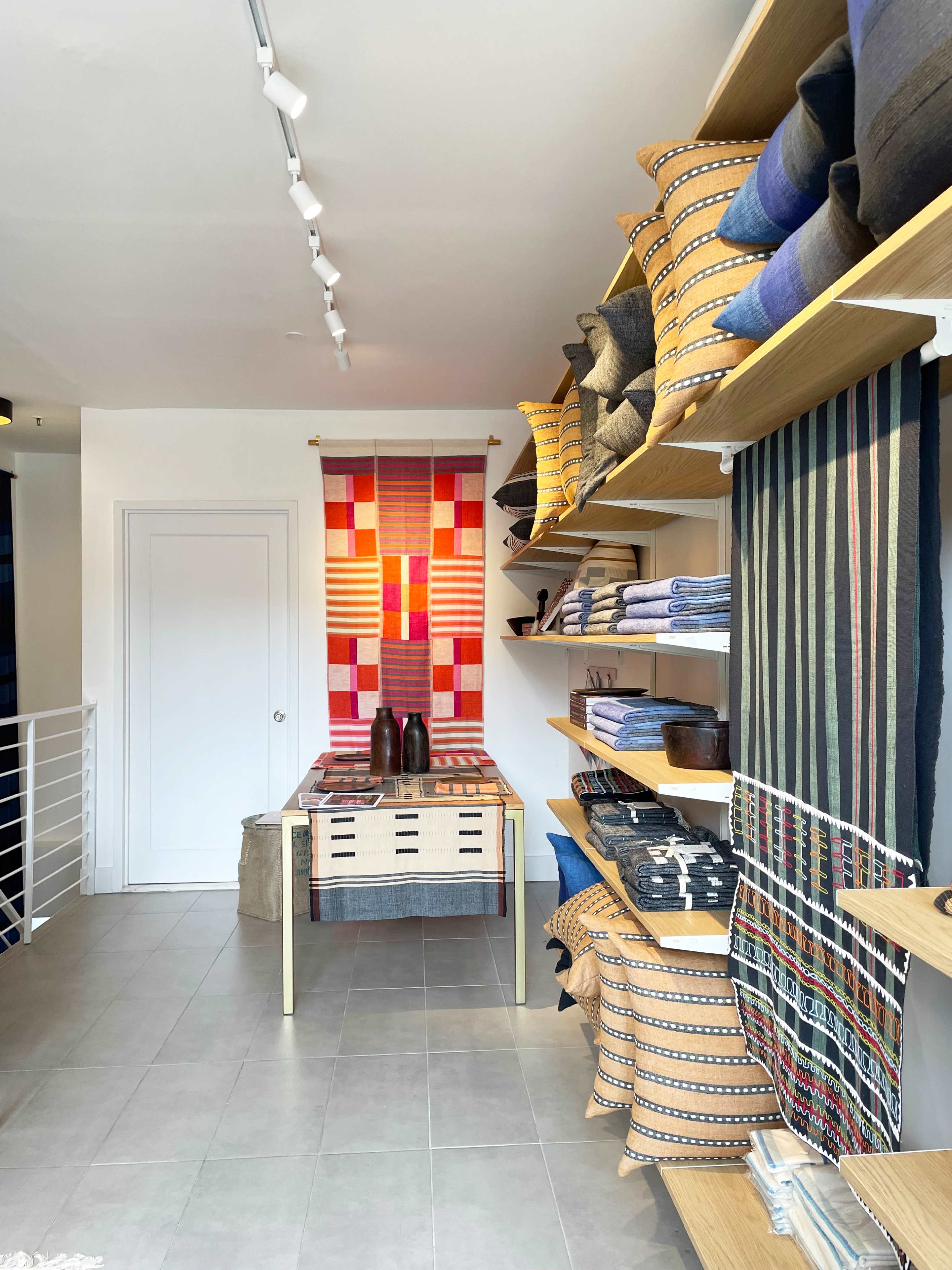 The image shows a well-organized interior of a store featuring shelves filled with colorful textiles and decorative items, including pillows and woven fabrics.