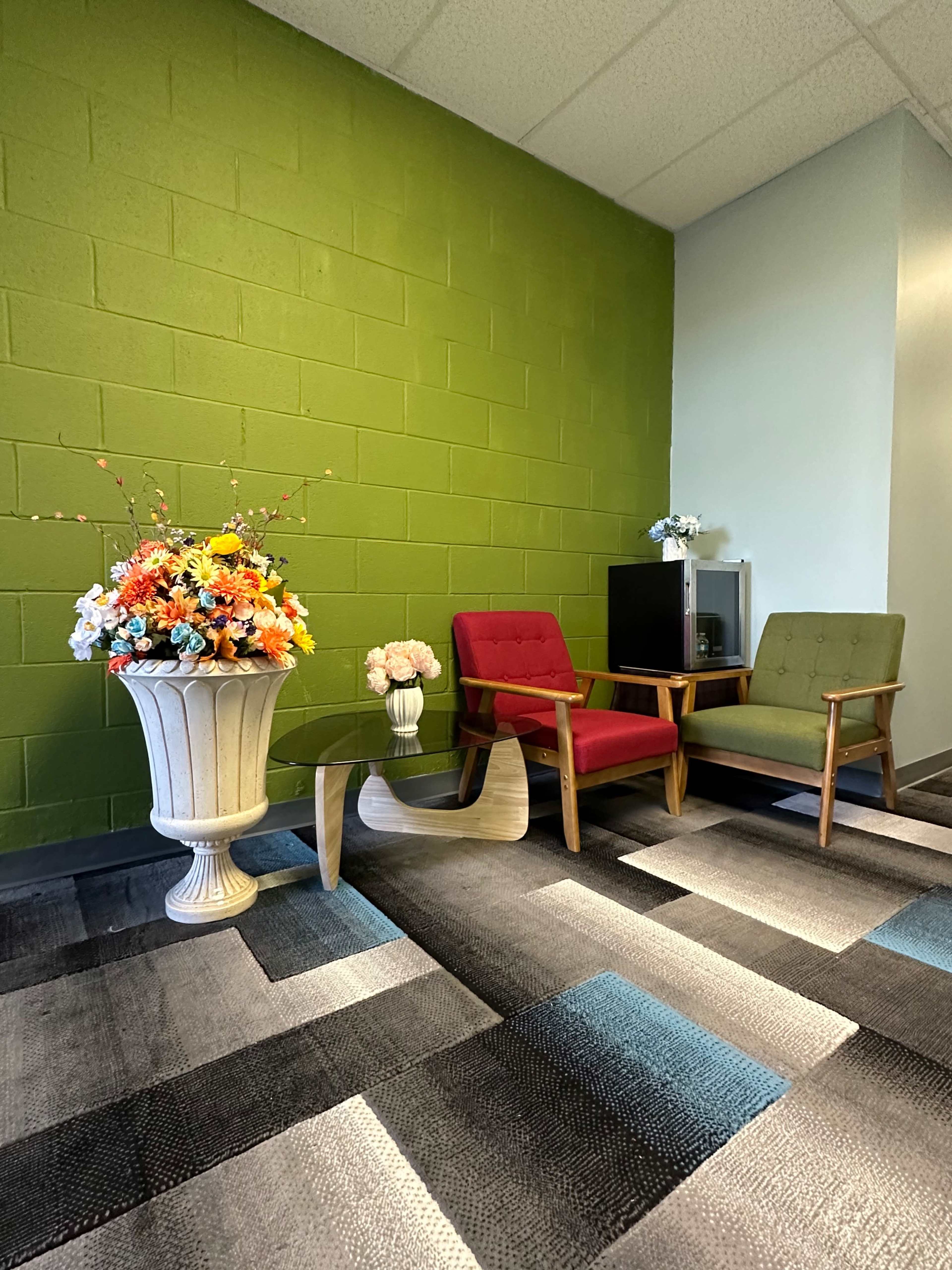 The image shows a corner of a room with a green wall, two chairs—one red and one green—and a decorative vase with artificial flowers beside a small glass table.