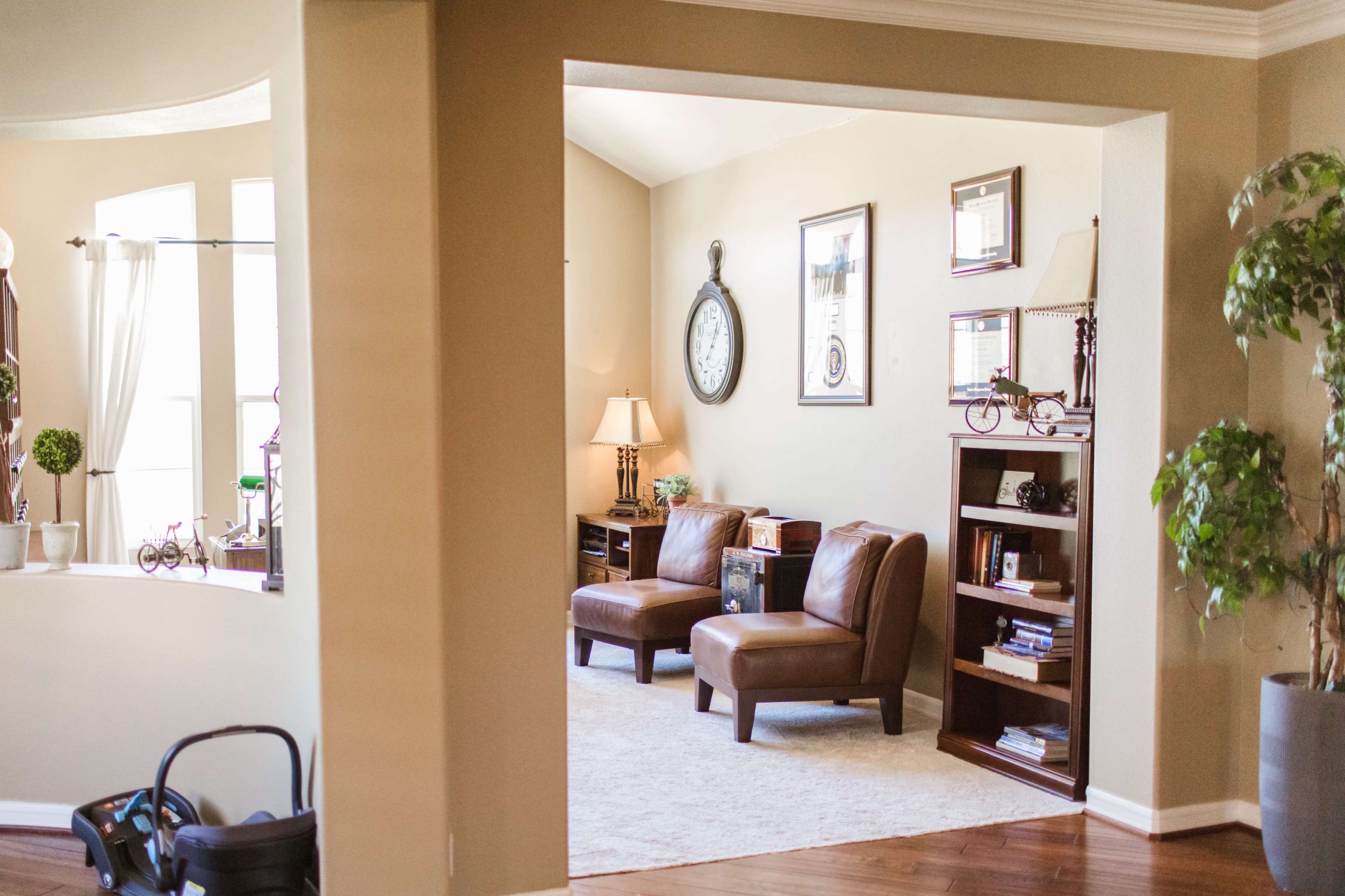 The image shows a living room with two brown armchairs, a lamp, a wall clock, and a bookshelf in an open, light-filled space.