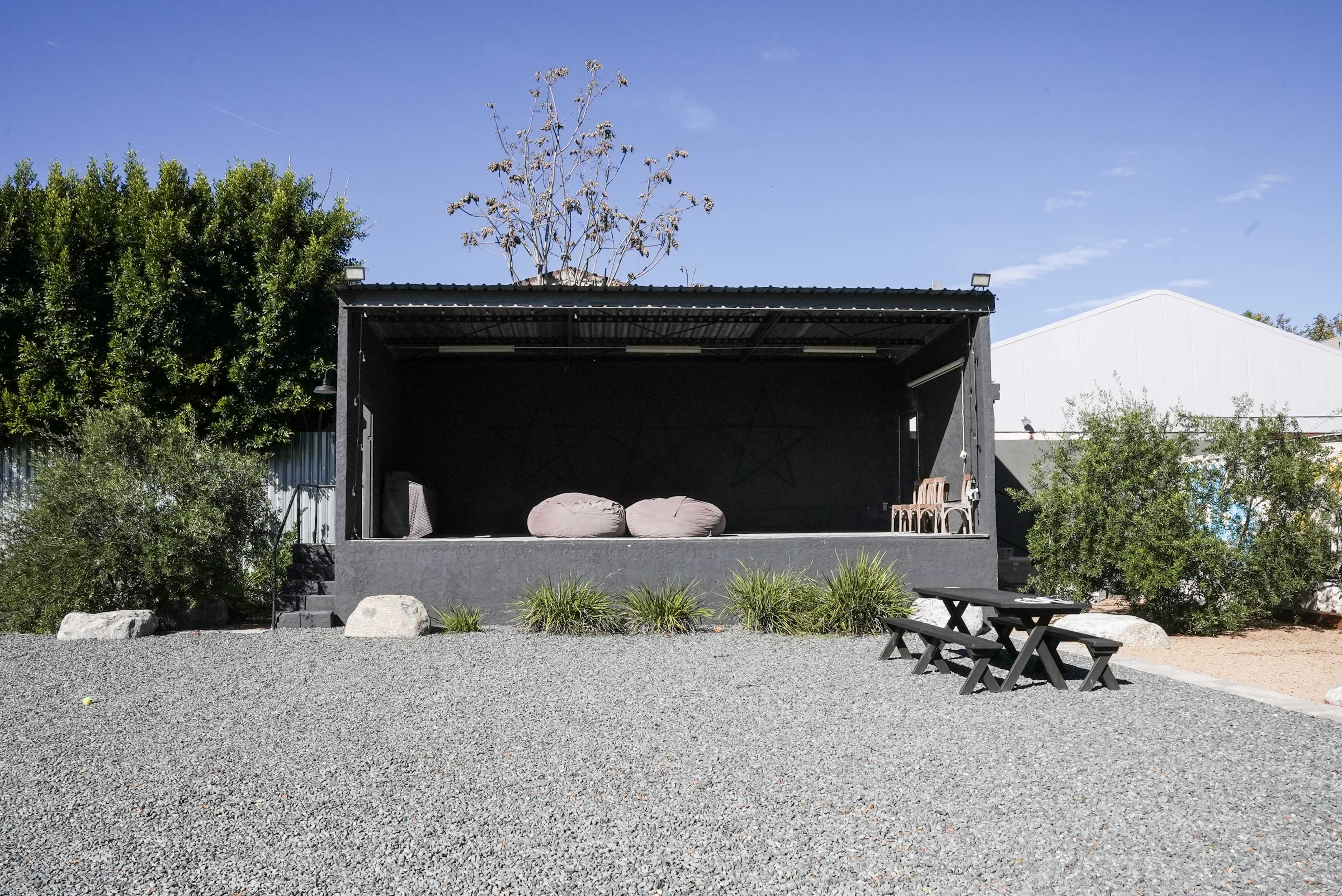 The image shows an outdoor stage area with two large bean bags, seating, and a gravel surface, surrounded by greenery and a building in the background.