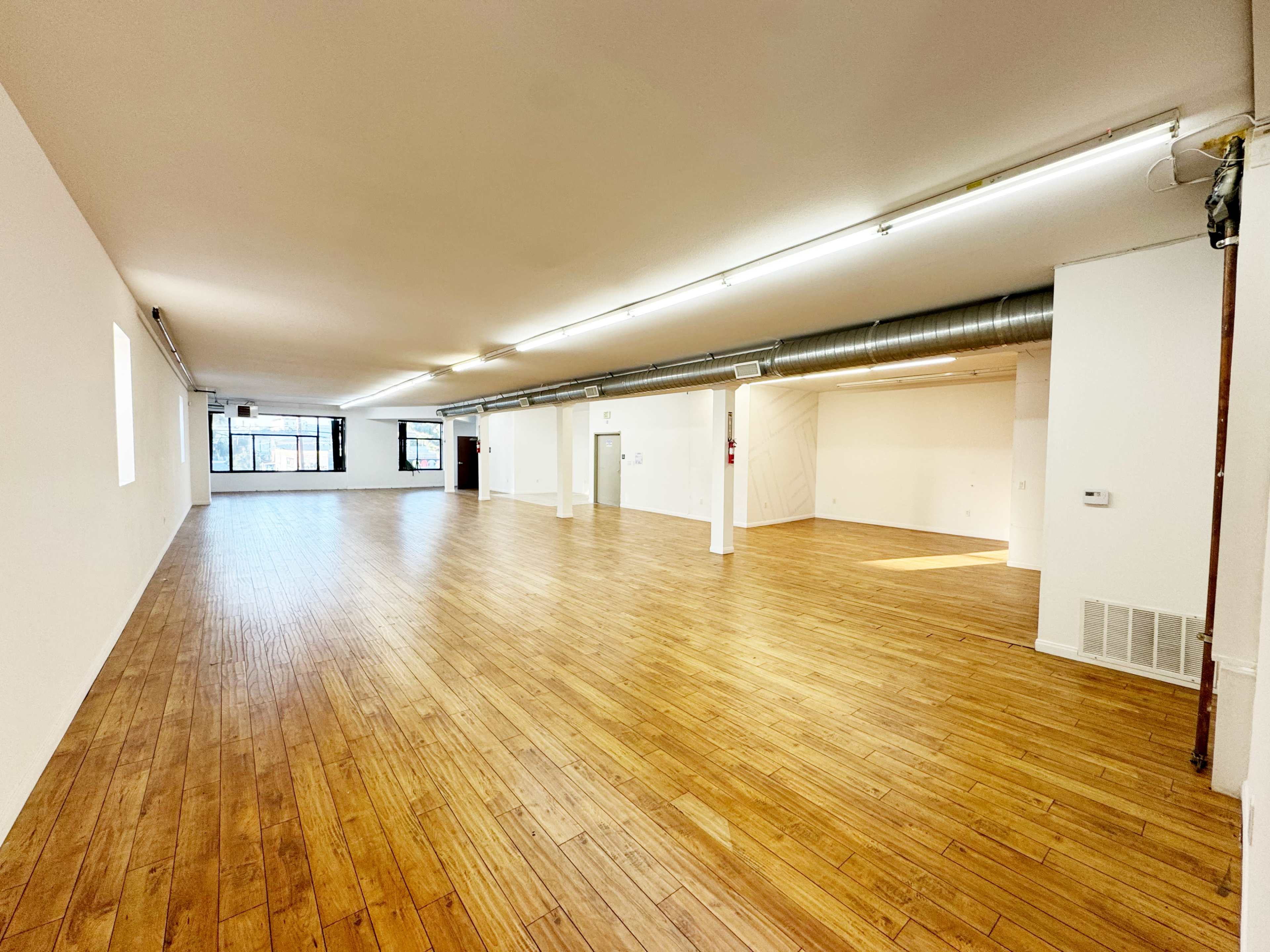 The image shows a spacious, empty room with wooden flooring and bright, natural light coming from windows on one side.