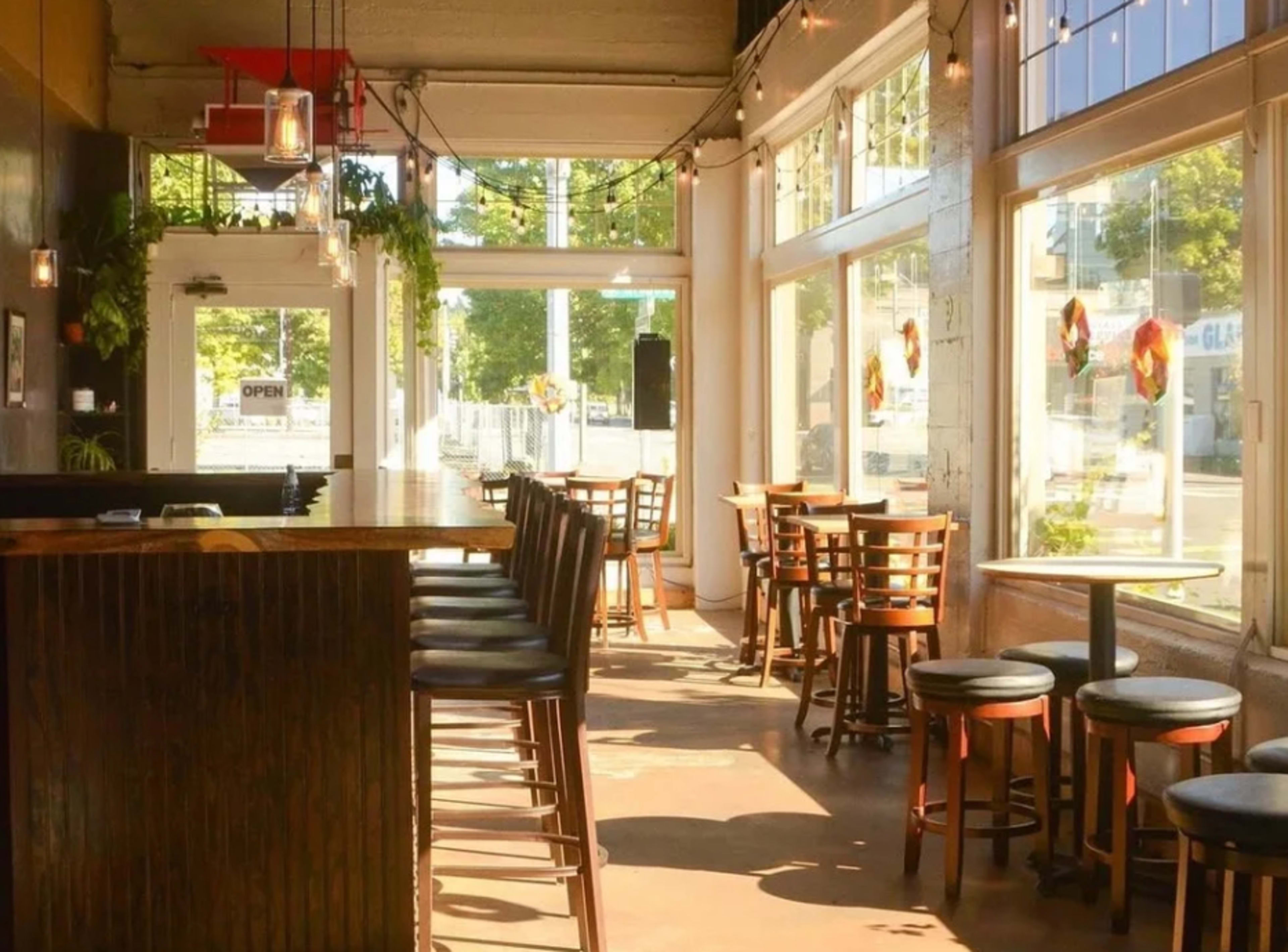 The image shows the interior of a cafe with wooden bar stools and tables arranged near large windows that allow natural light to fill the space.