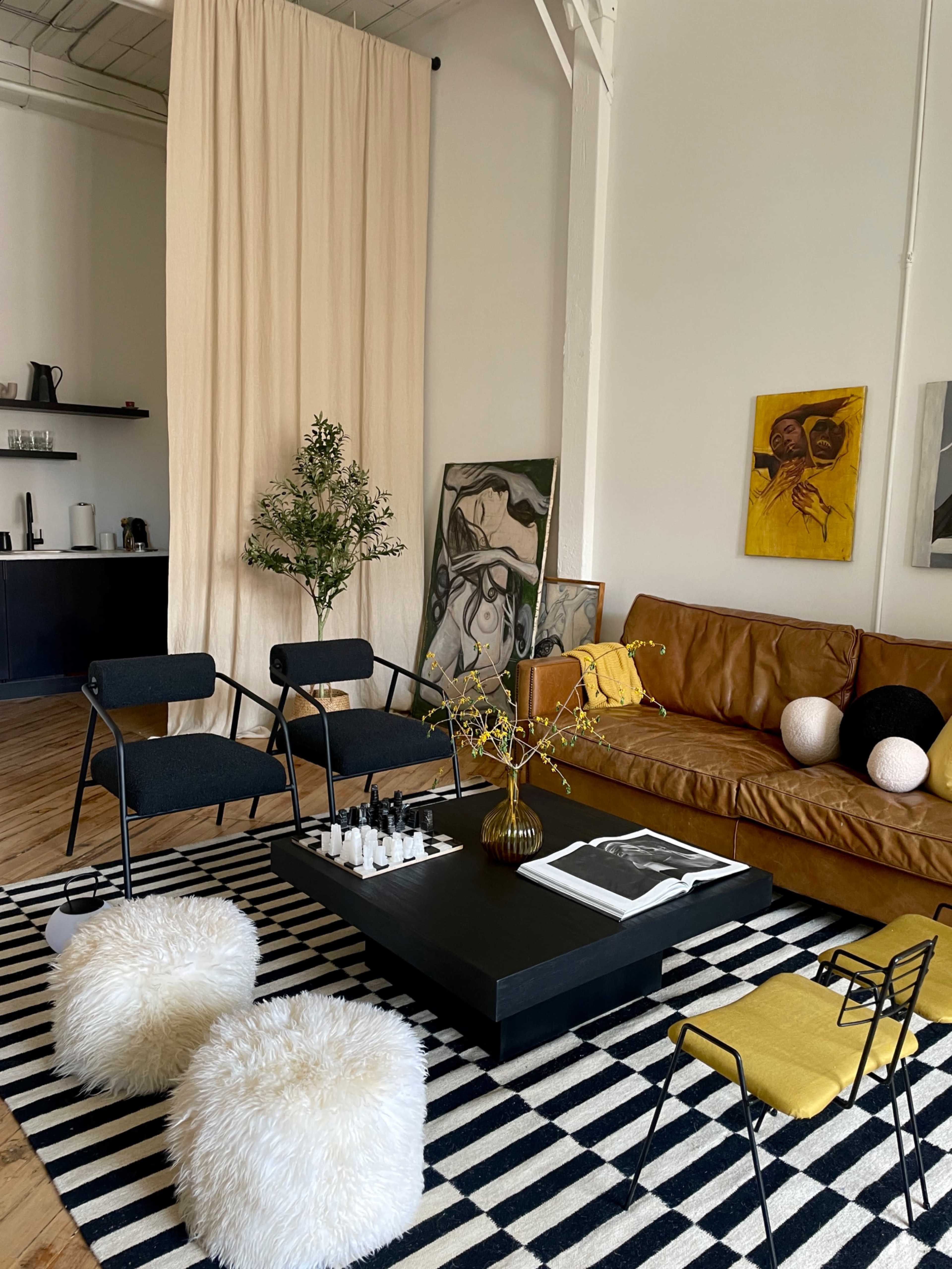 The image shows a modern living room with a brown leather sofa, black mid-century chairs, a striped rug, a black coffee table, and decorative plants and artwork on the walls.