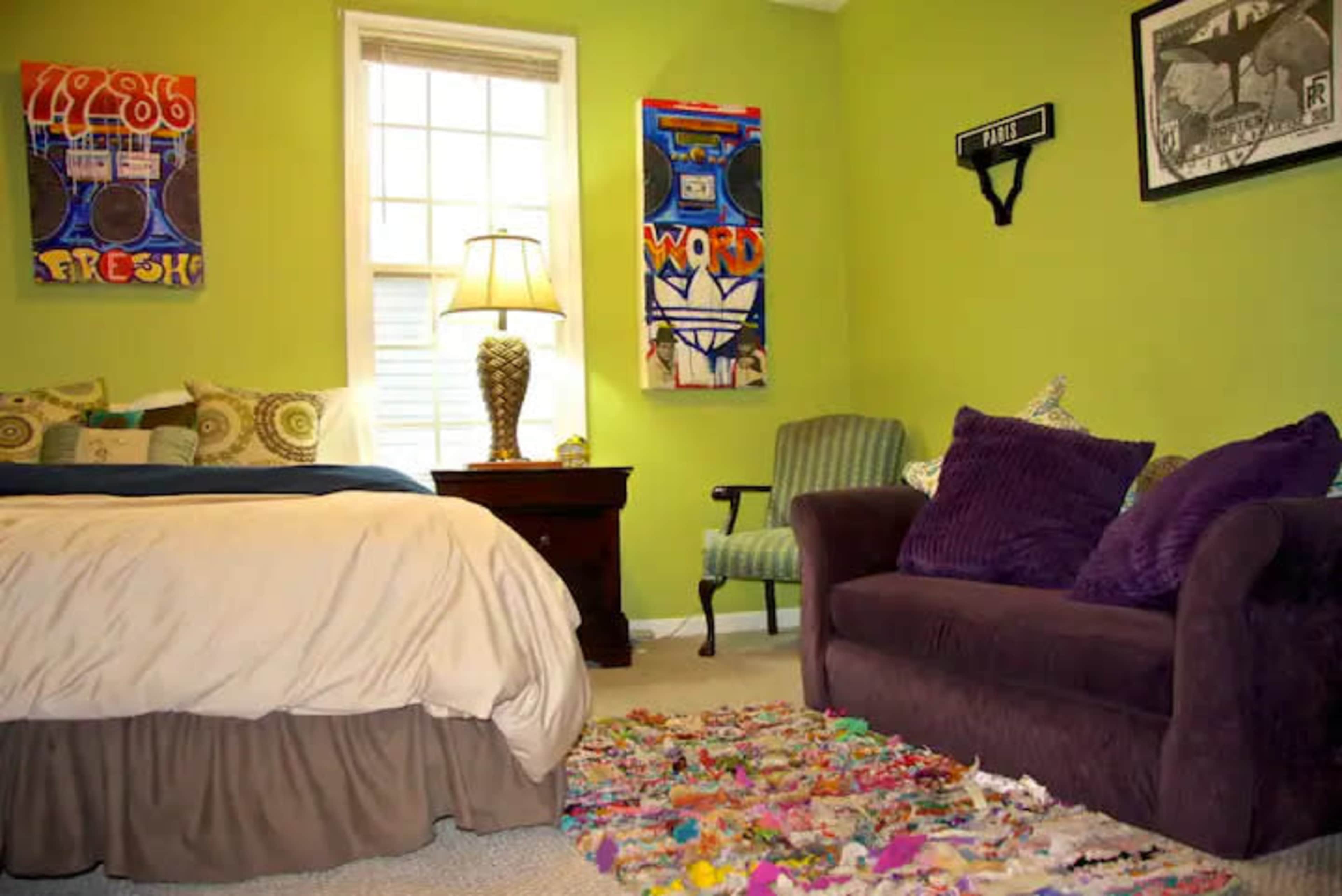 The room features a bed with a light-colored comforter, a small table with a lamp, a green wall adorned with artwork, and a purple couch beside a colorful rug.