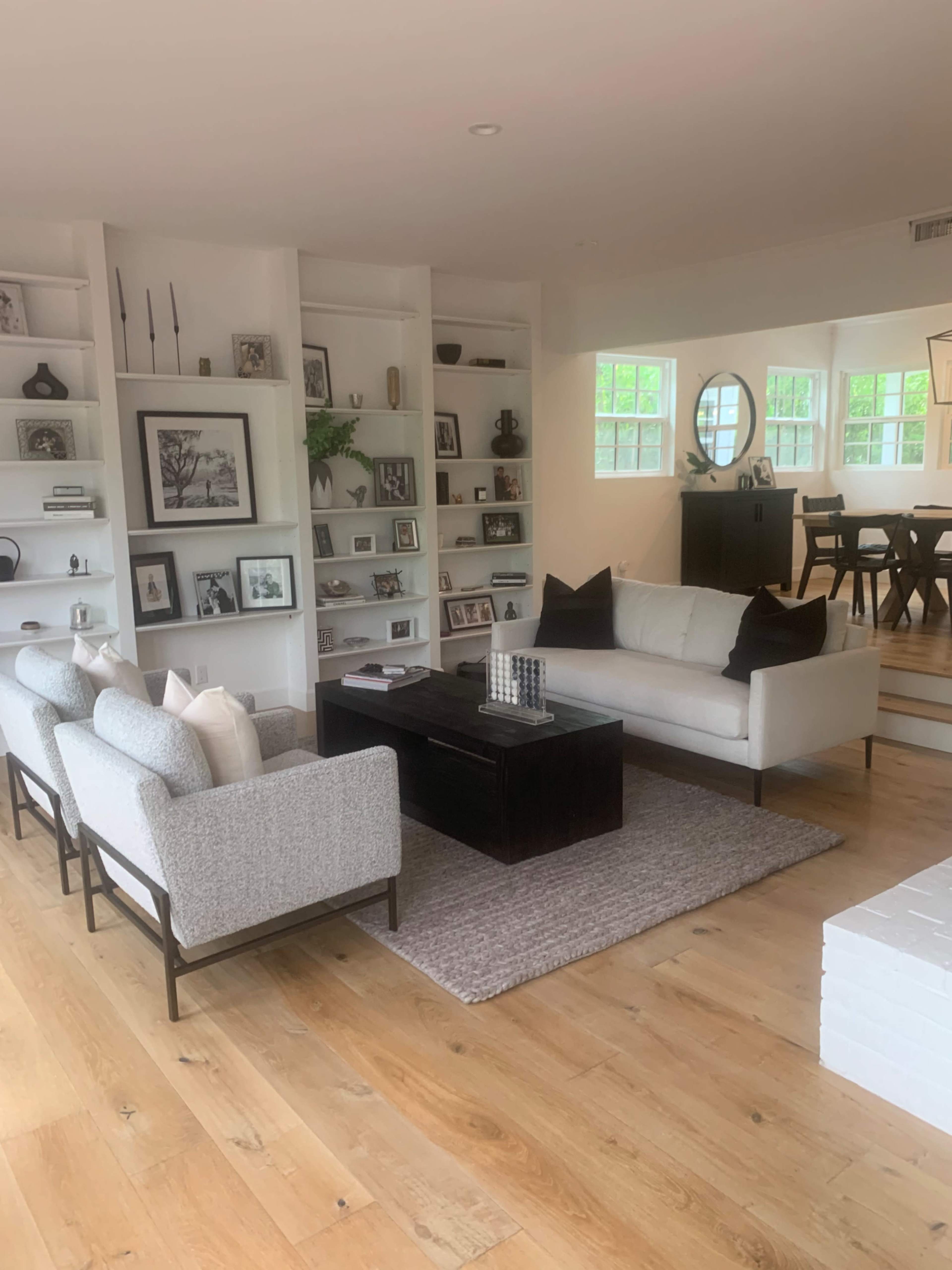 The image shows a living room with a sofa and two armchairs around a coffee table, surrounded by built-in shelves filled with decorative items, and a dining area visible in the background.