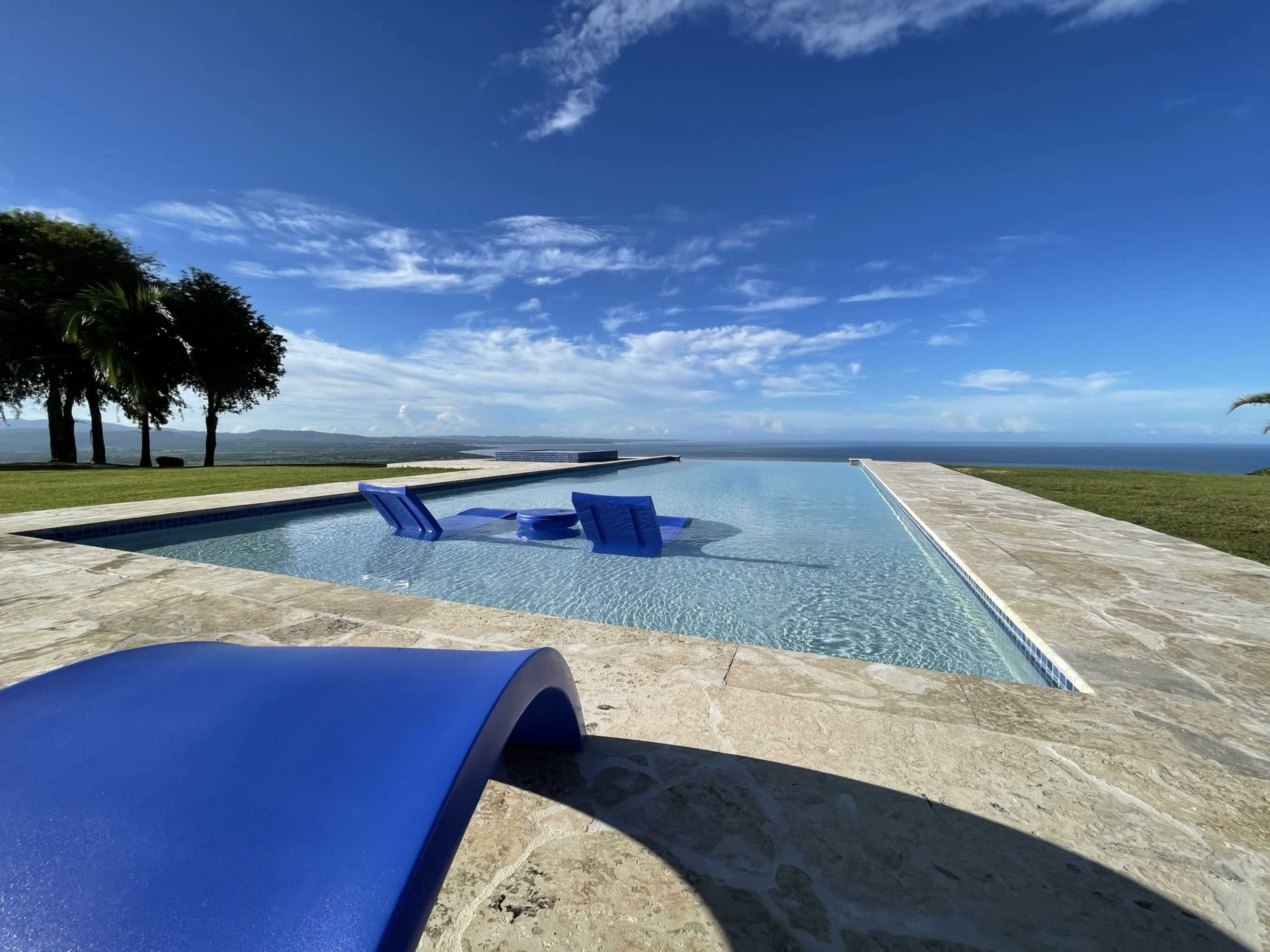 A long rectangular swimming pool with blue lounge chairs is set against a backdrop of palm trees and a clear sky over a distant body of water.