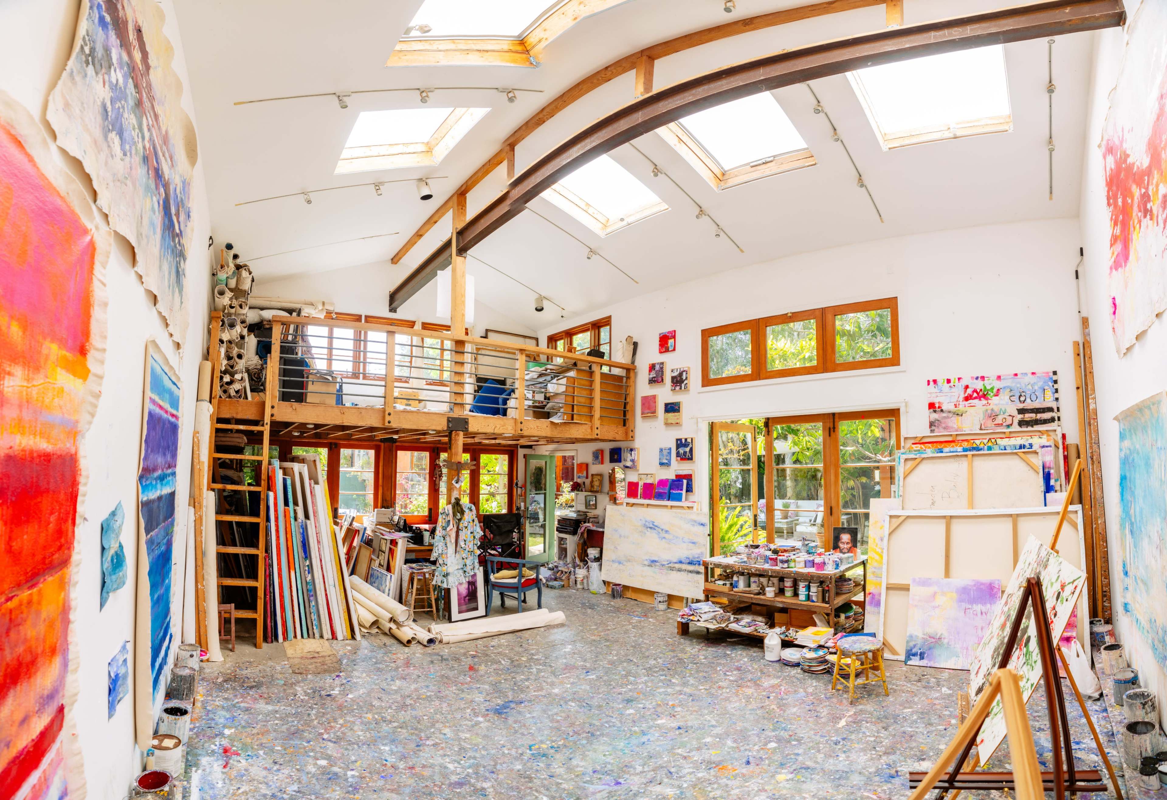 A spacious artist's studio with large windows, a lofted area, and colorful paintings displayed on the walls and floor.
