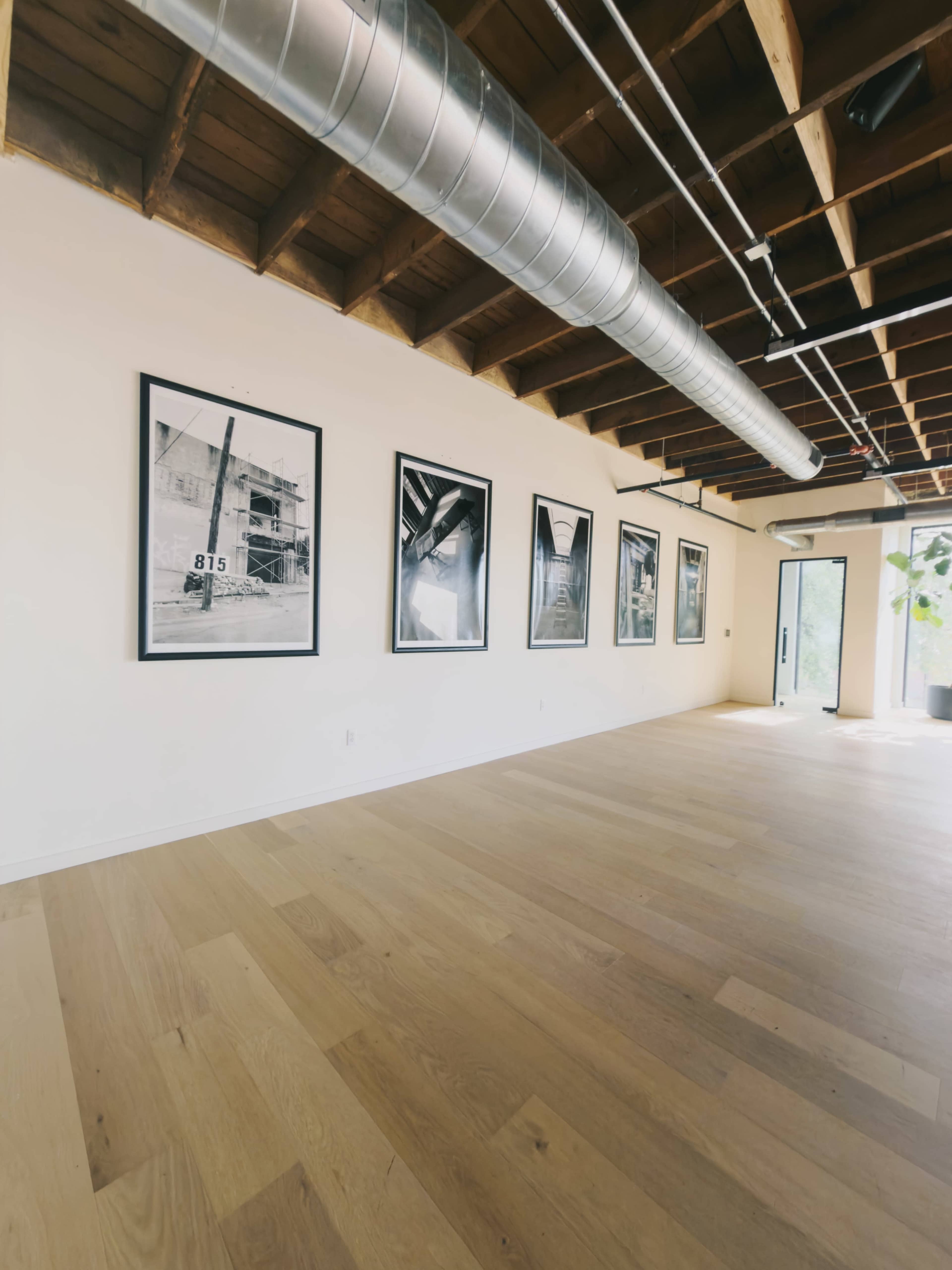 The image shows a spacious room with wooden flooring, featuring several black-and-white framed photographs on the walls and visible industrial-style ductwork on the ceiling.