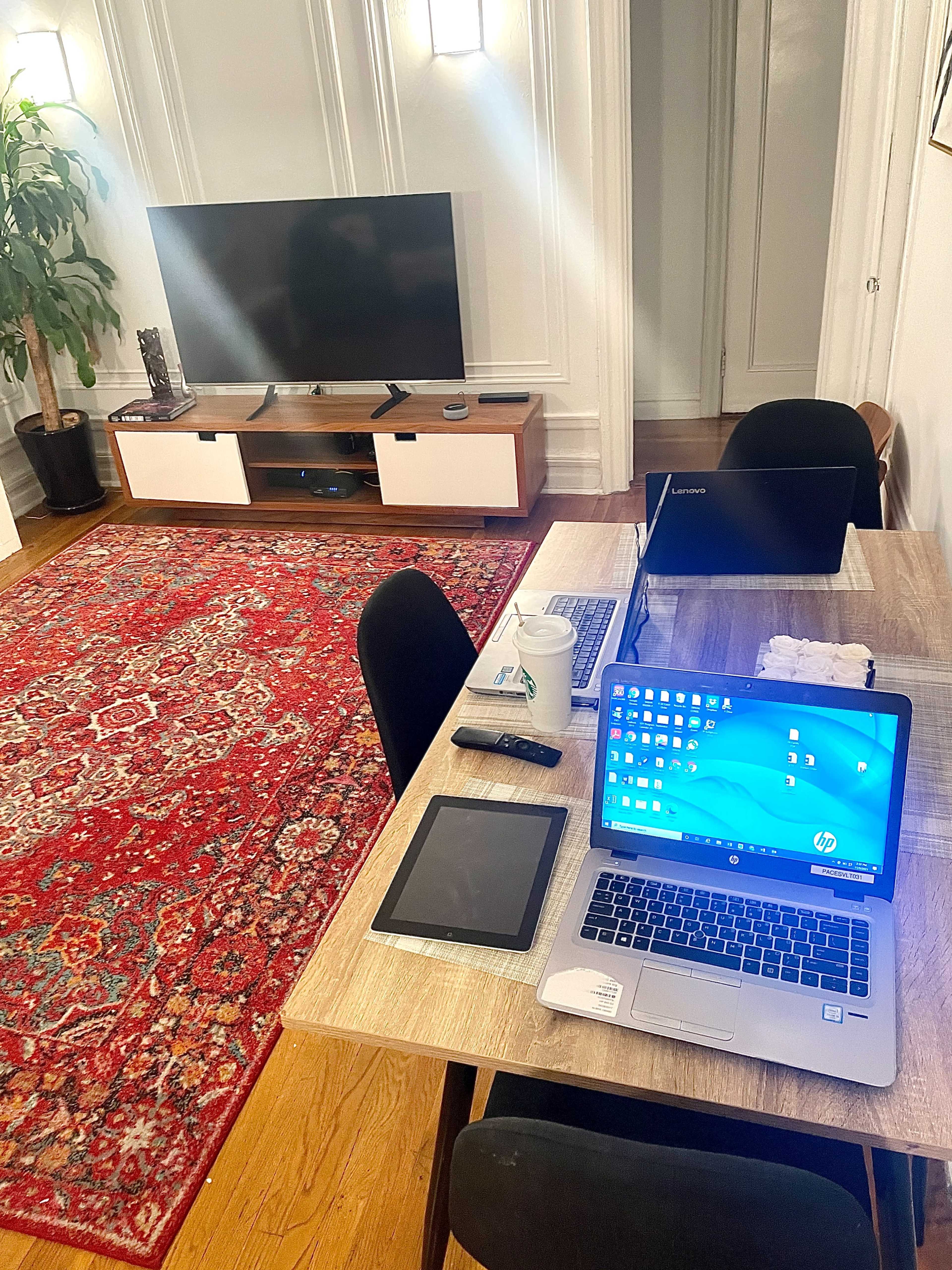 A modern workspace features a wooden table with a laptop, tablet, and accessories, set against a backdrop of a television and a large area rug.