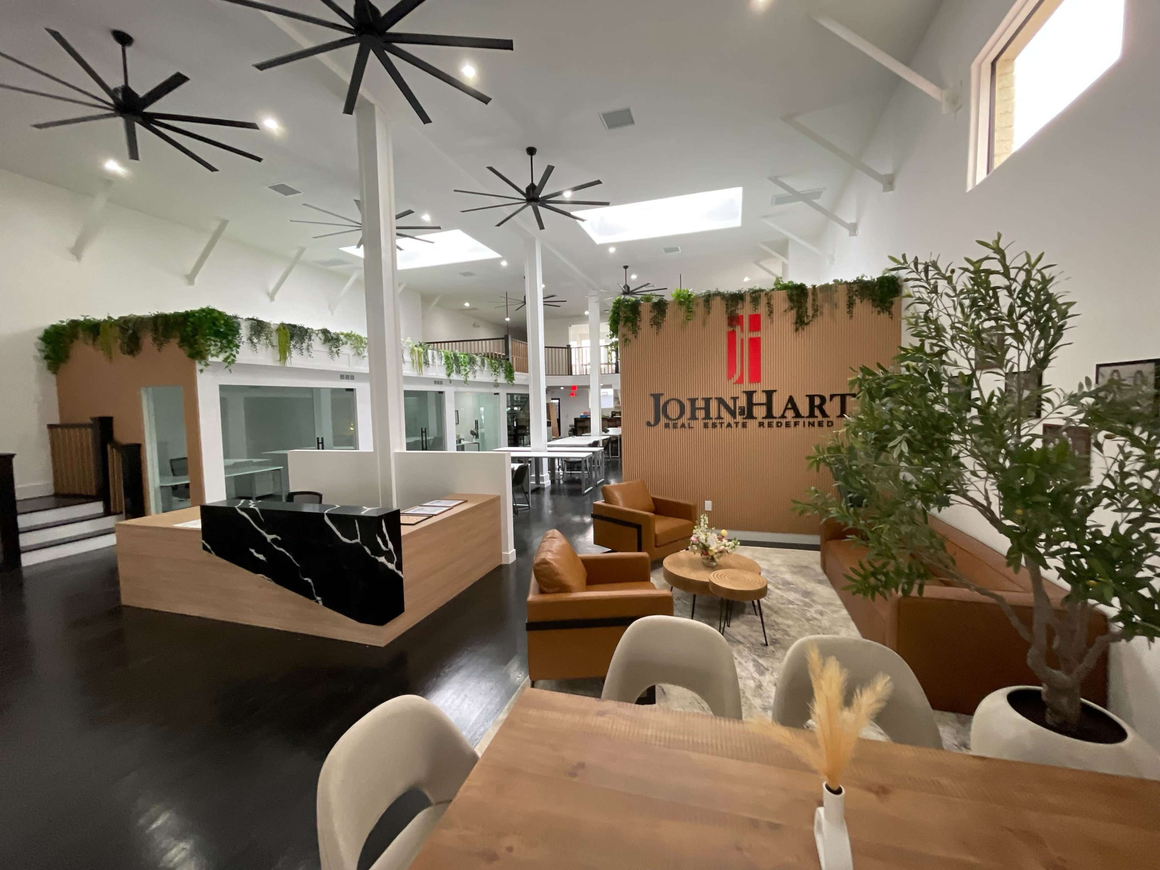 The interior of a modern real estate office features a reception area with a large logo, seating arrangements, and abundant natural light from overhead windows.