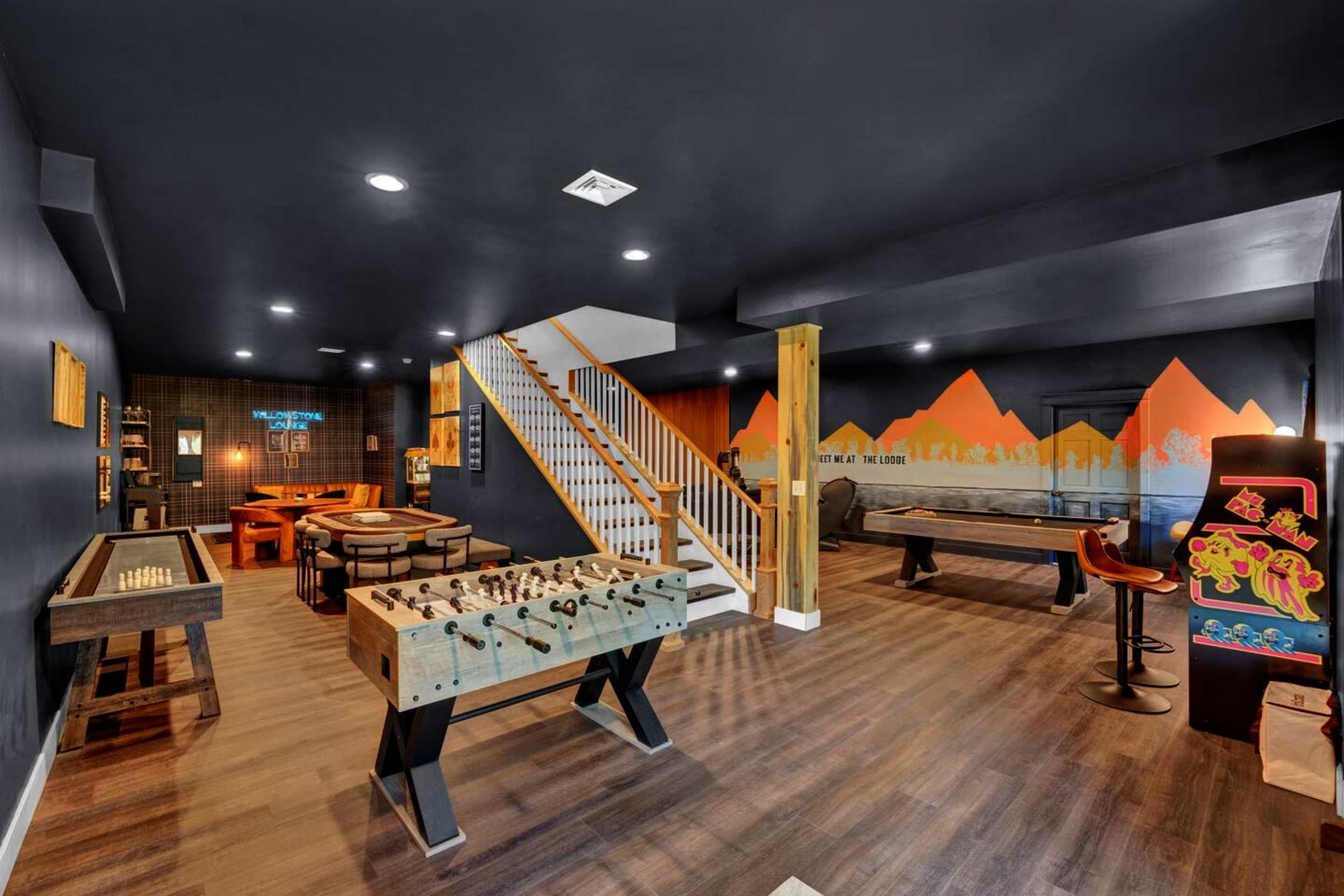 The image shows a basement game room featuring a foosball table, a pool table, a chess set, and a colorful wall mural of mountains.