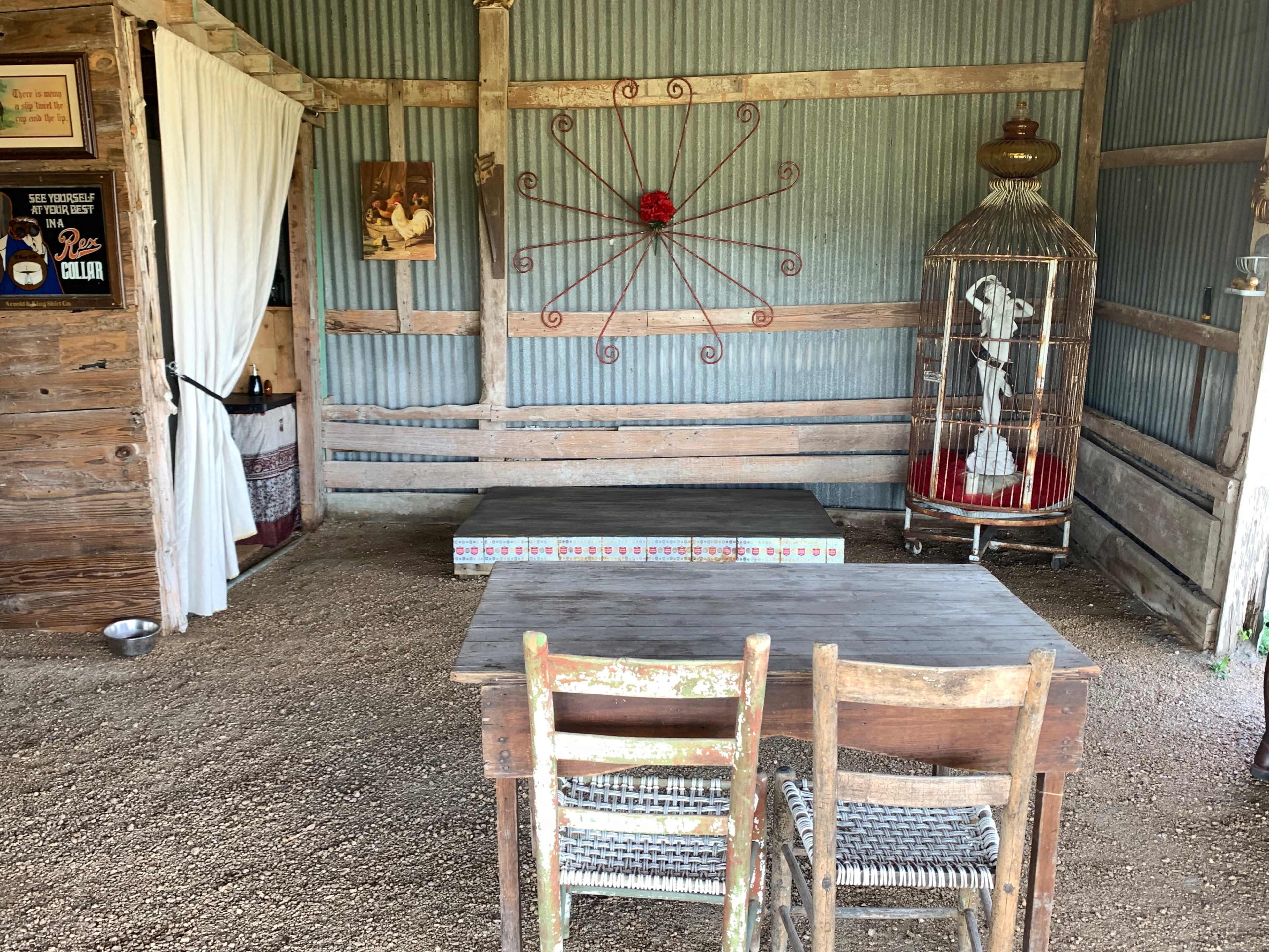 The image shows an interior space with wooden walls, a small table, two wooden chairs, and a decorative birdcage against a backdrop of corrugated metal.