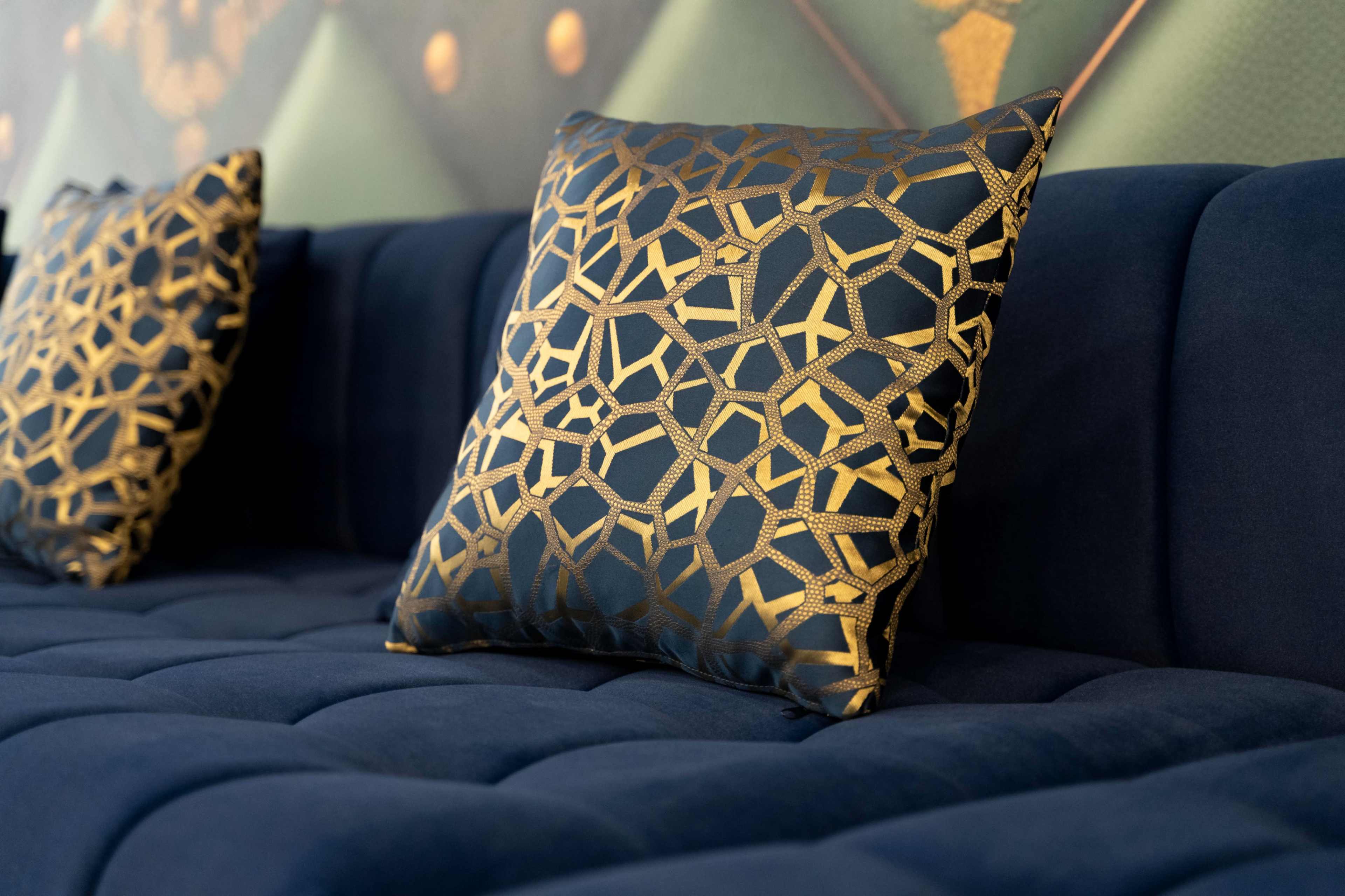 The image shows a navy blue tufted couch adorned with two decorative pillows featuring a geometric gold pattern.