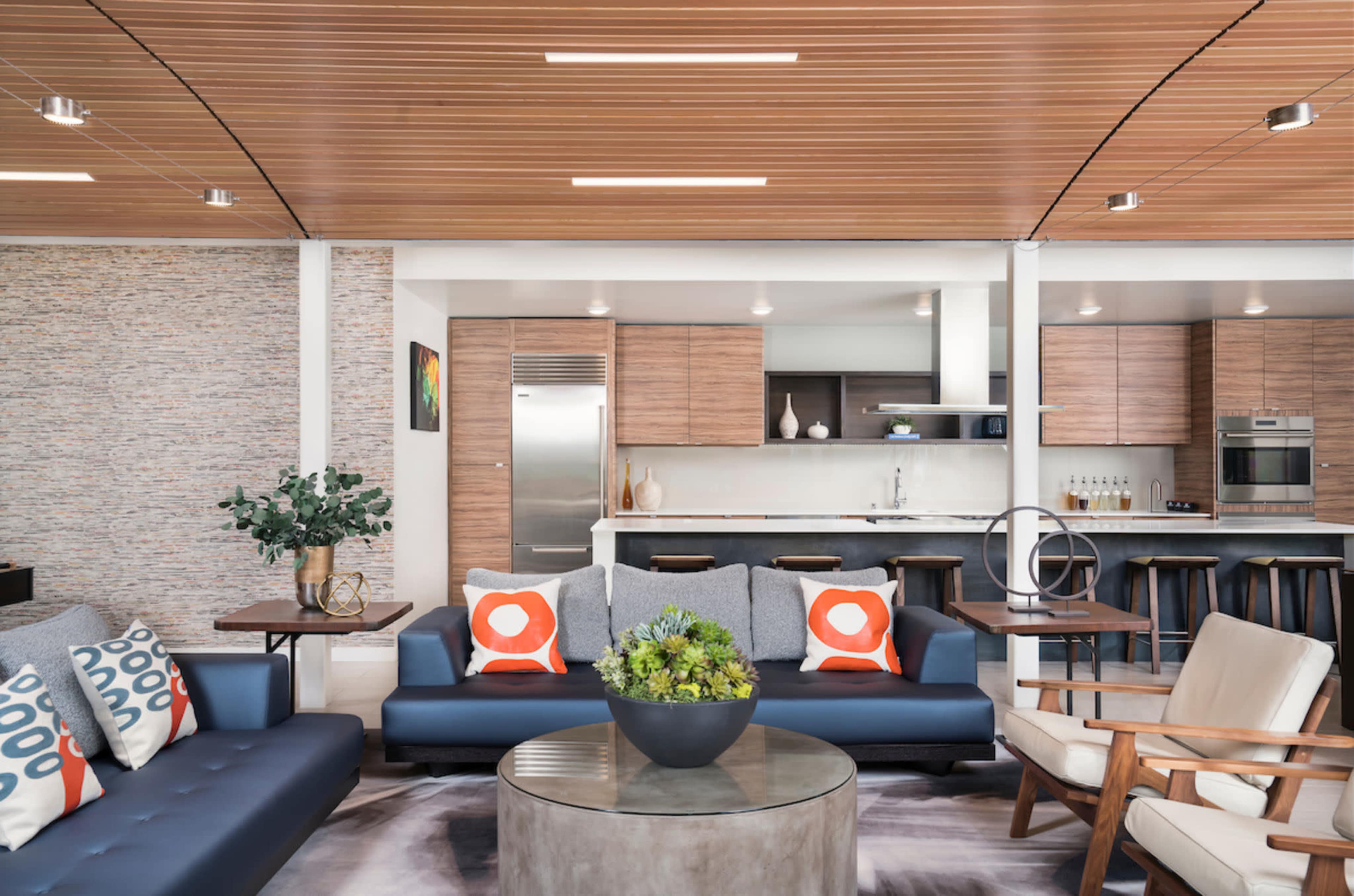 A modern living area with two sofas, a coffee table, and a kitchen in the background featuring wooden cabinetry and stainless steel appliances.