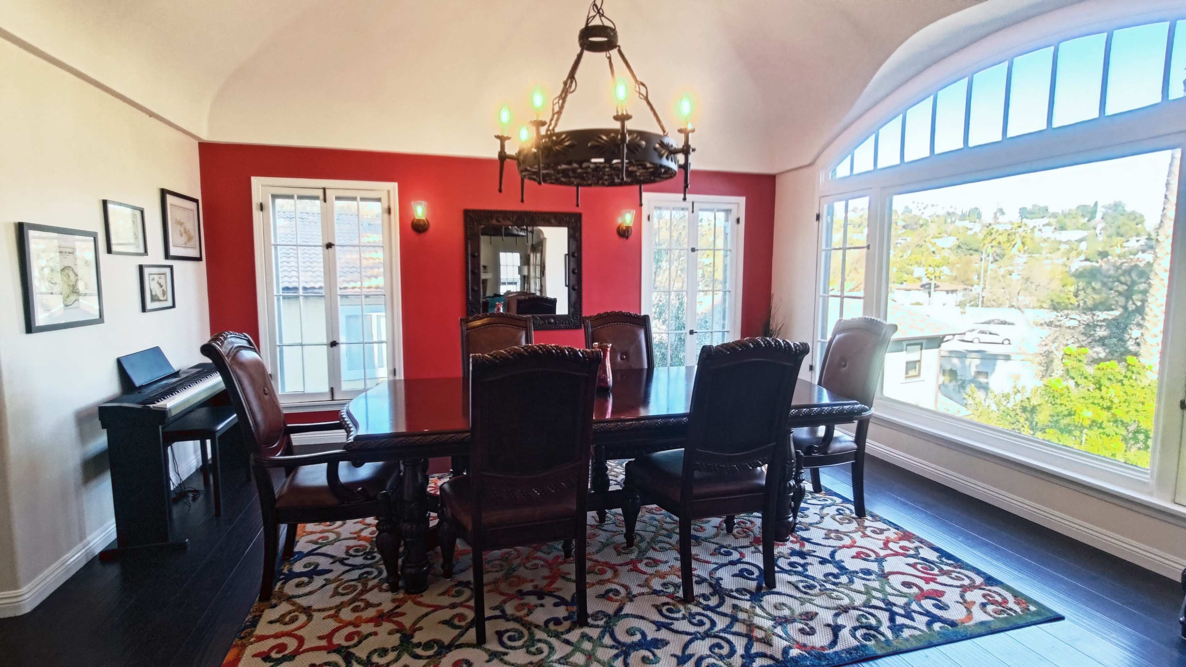 The dining room features a large wooden table surrounded by chairs, set against a backdrop of red walls and large windows that allow natural light to enter.