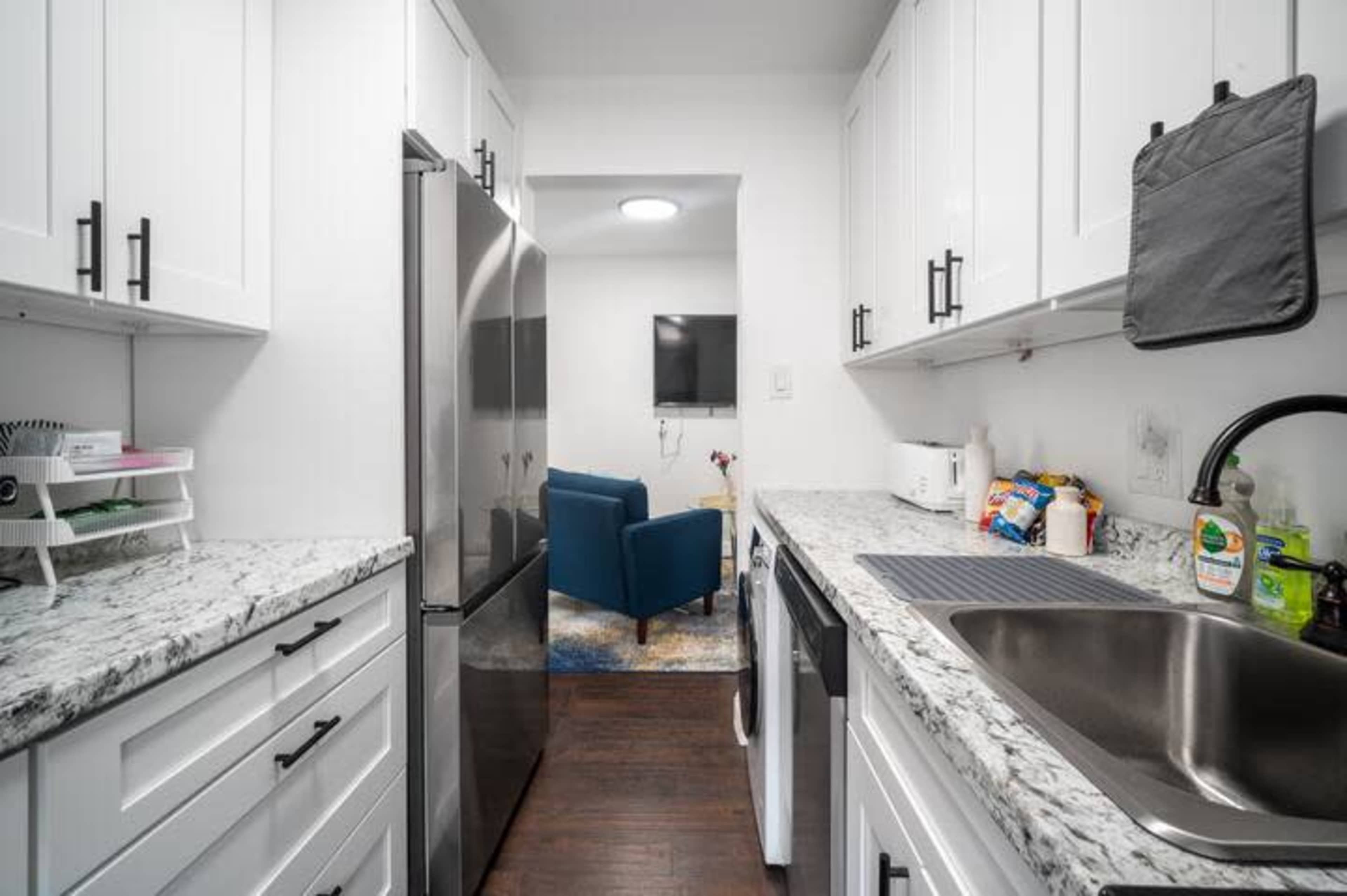The image shows a narrow kitchen with white cabinets, stainless steel appliances, and a view into a living area with a blue couch.