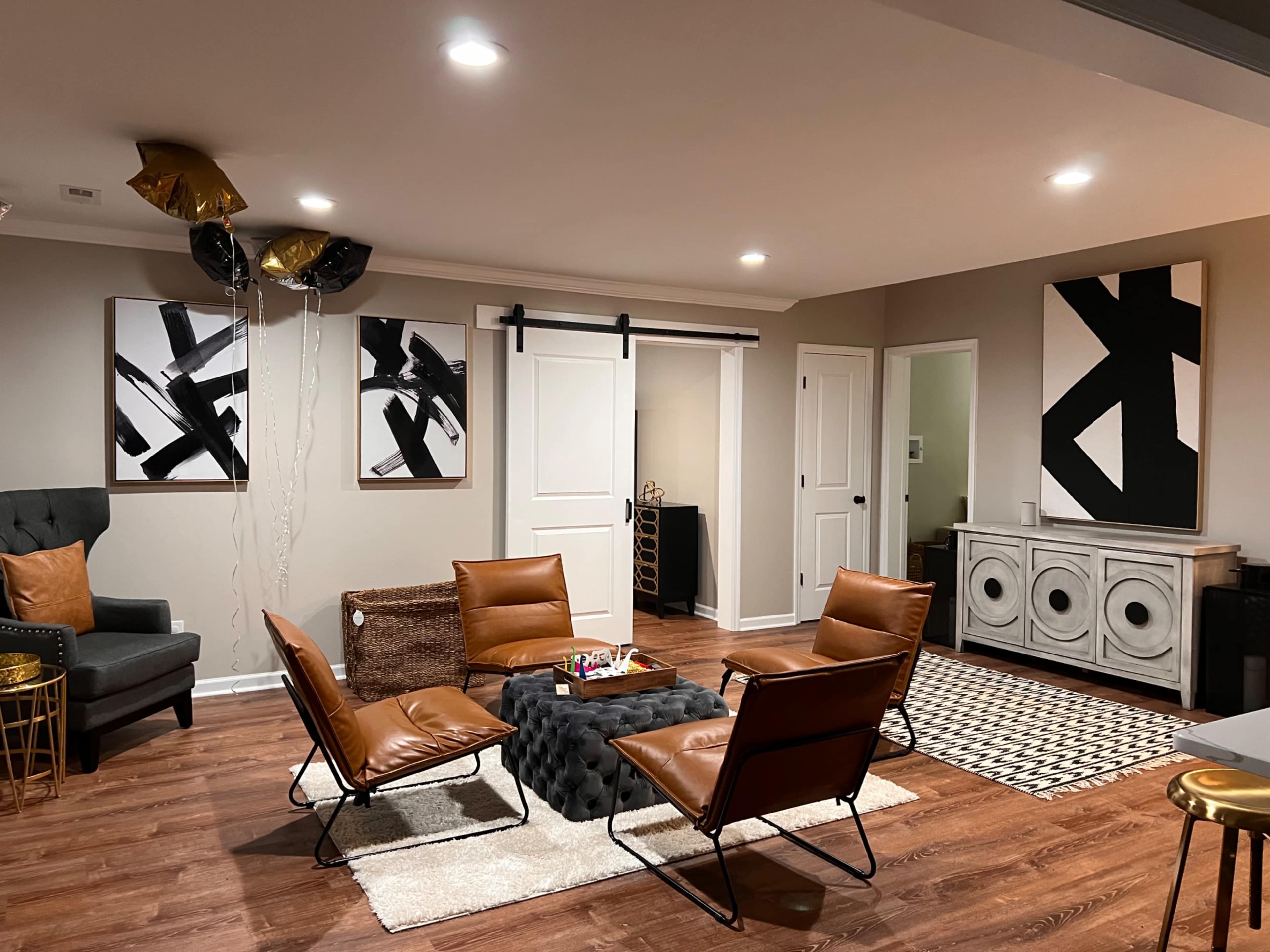 A modern living room with brown leather chairs arranged around a black coffee table, featuring abstract black-and-white art on the walls and decorative balloons in one corner.