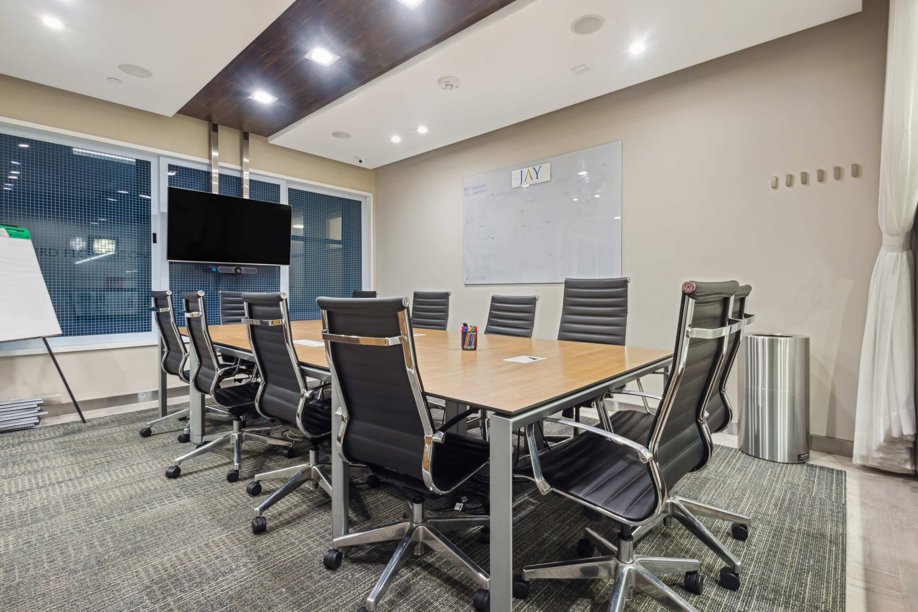 A modern conference room features a large wooden table surrounded by eight black office chairs, with a wall-mounted TV and a whiteboard in the background.