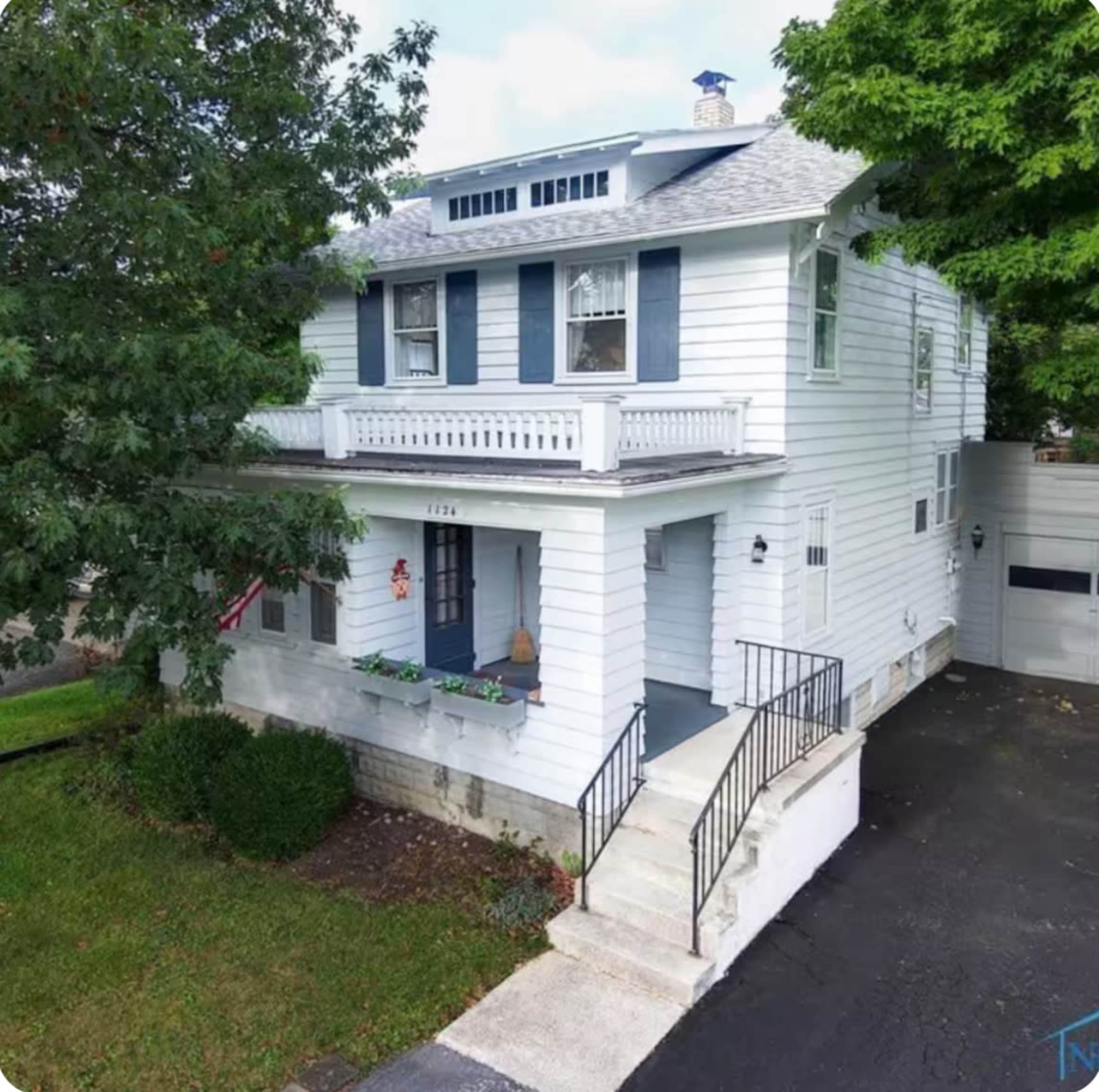 The image shows a two-story white house with blue shutters and a front porch, surrounded by greenery and parking space.