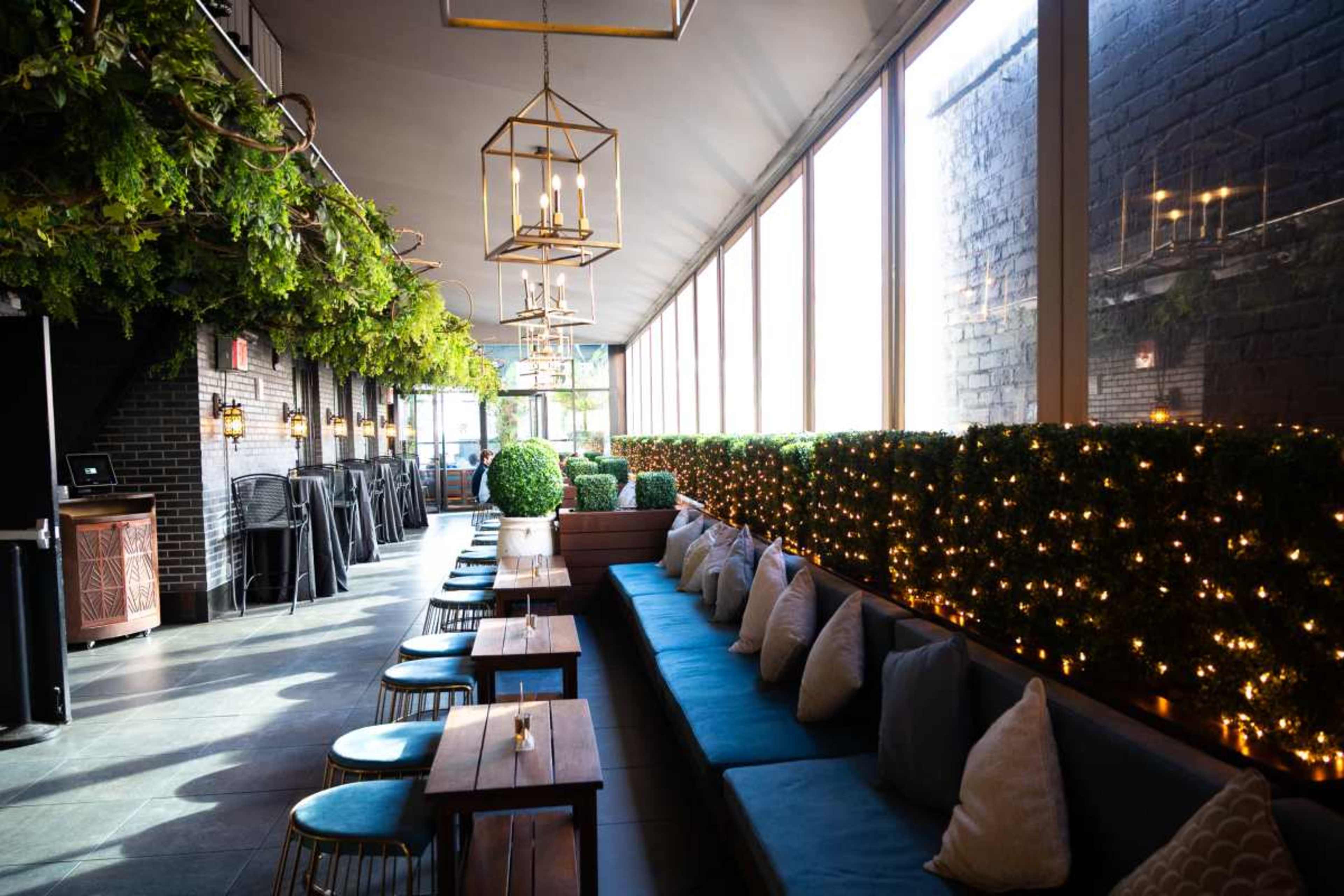 The image shows a beautifully designed indoor space with long benches and tables, surrounded by lush greenery and warm lighting.