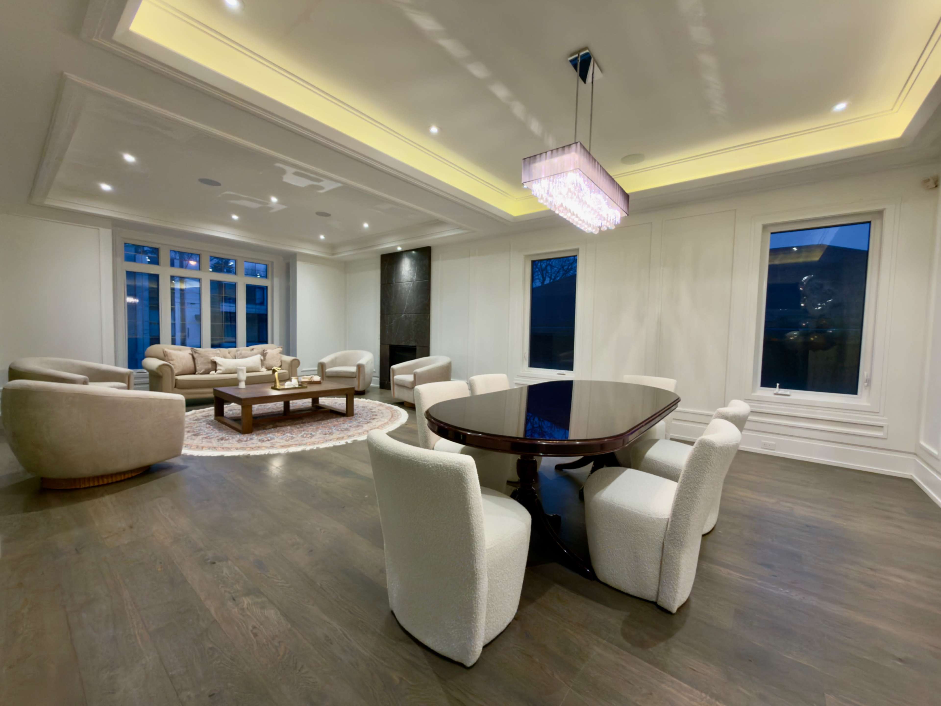 The image shows a modern, elegantly designed living space featuring a dining table surrounded by plush chairs, a cozy seating area with a sofa, and large windows allowing natural light to enter.